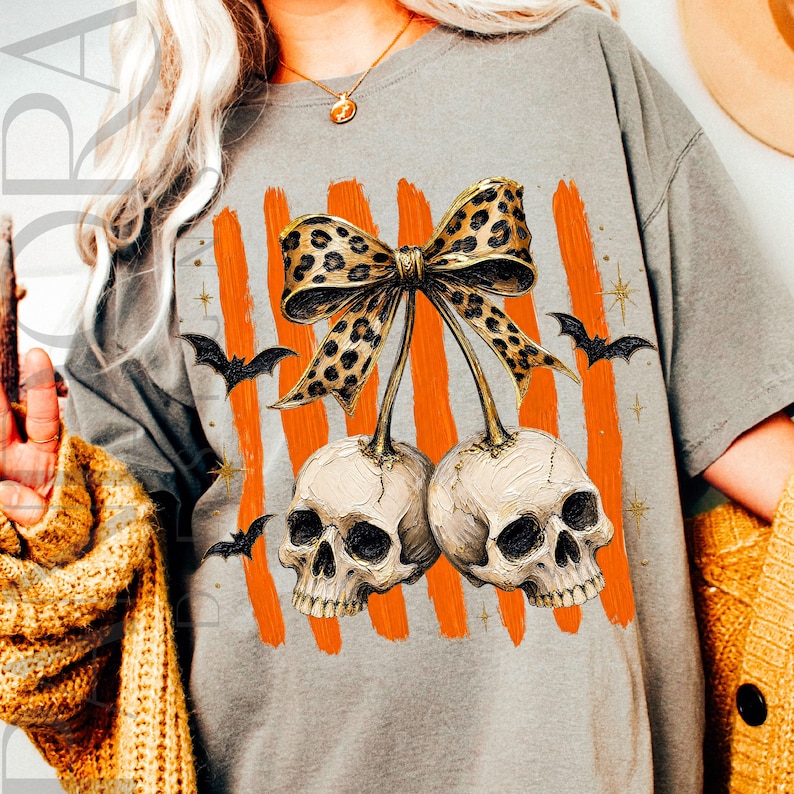 May include: A grey t-shirt featuring a graphic of two human skulls connected by stems to a large leopard print bow. The design includes vertical orange brushstroke stripes in the background, small black bats, and gold star accents.