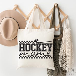 May include: White canvas tote bag with black text that reads "Hockey Mom" with a small heart symbol. The text is in a bold, blocky font. The bag is hanging on a wooden rack with a brown hat and a beige mesh bag.
