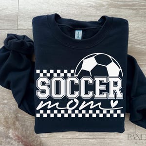 Pu&ograve; includere: Felpa nera con una grafica di pallone da calcio bianco e il testo "Soccer Mom" con un cuore.