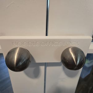 May include: White door lock with two brushed metal knobs. The text "NOODLE OFFICE LOCK" is printed on the white plastic. The lock is designed to secure a door.