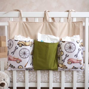 Könnte beinhalten: Ein beiger Stoff-Organizer für das Kinderbett mit drei Taschen. Zwei Taschen zeigen einen Vintage-Auto- und Werkzeugdruck, während die mittlere Tasche einfarbig grün ist. Der Organizer hängt an der Krippenschiene.