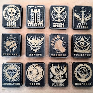 Könnte beinhalten: Eine Sammlung von zwölf rechteckigen Spielmarken aus Holz mit schwarzen Hintergründen und goldfarbenen eingravierten Designs und Texten. Die Marken zeigen verschiedene Symbole und Wörter wie "Death Touch", "Defender" und "Double Strike".