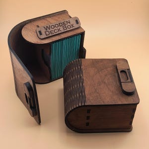 Peut inclure: Boîte de jeu en bois marron foncé avec l'inscription "WOODEN DECK BOX". La boîte est ouverte, révélant une pile de cartes turquoise. La boîte a un design incurvé et est en bois.