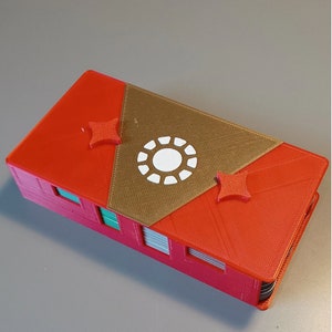 Puede incluir: Una caja roja y dorada impresa en 3D con un dise&ntilde;o de c&iacute;rculo blanco en la parte superior. La caja tiene dos acentos rojos en forma de estrella en la parte superior.