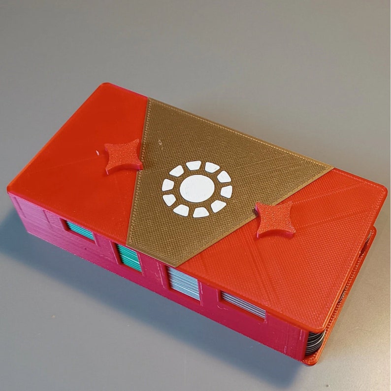 Puede incluir: Una caja roja y dorada impresa en 3D con un dise&ntilde;o de c&iacute;rculo blanco en la parte superior. La caja tiene dos acentos rojos en forma de estrella en la parte superior.