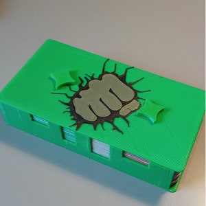 Puede incluir: Una caja de cartas verde impresa en 3D con un gr&aacute;fico de pu&ntilde;o negro y gris. El pu&ntilde;o est&aacute; rompiendo la superficie de la caja. Hay dos peque&ntilde;as estrellas negras y grises en la caja.