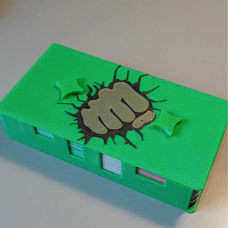 Puede incluir: Una caja de cartas verde impresa en 3D con un gr&aacute;fico de pu&ntilde;o negro y gris. El pu&ntilde;o est&aacute; rompiendo la superficie de la caja. Hay dos peque&ntilde;as estrellas negras y grises en la caja.