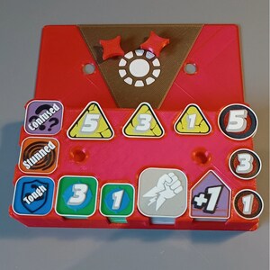 Puede incluir: Una bandeja de juego de pl&aacute;stico rojo con una parte superior marr&oacute;n. La bandeja tiene ranuras para guardar varias piezas de juego, incluidos dados, fichas y cartas. La bandeja tambi&eacute;n tiene un peque&ntilde;o c&iacute;rculo blanco con un contorno negro en el centro de la parte superior. La bandeja est&aacute; etiquetada con las palabras "Confused", "Stunned", "Tough", "3", "1", "5", "3", "1", "5", "3", "1", "1", y "+1".