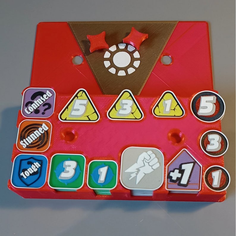 Puede incluir: Una bandeja de juego de pl&aacute;stico rojo con una parte superior marr&oacute;n. La bandeja tiene ranuras para guardar varias piezas de juego, incluidos dados, fichas y cartas. La bandeja tambi&eacute;n tiene un peque&ntilde;o c&iacute;rculo blanco con un contorno negro en el centro de la parte superior. La bandeja est&aacute; etiquetada con las palabras "Confused", "Stunned", "Tough", "3", "1", "5", "3", "1", "5", "3", "1", "1", y "+1".