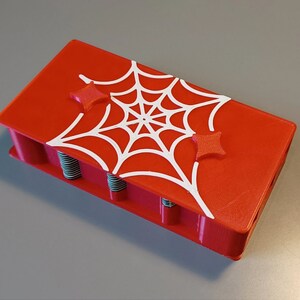 Puede incluir: Una caja roja impresa en 3D con un dise&ntilde;o de telara&ntilde;a blanca y dos botones rojos en forma de estrella. La caja tiene una serie de crestas a lo largo de la parte inferior.