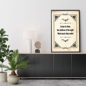 May include: A framed print with a vintage parchment background and black decorative borders. The text reads, "Listen to them, the children of the night. What music they make!" Two bat illustrations are above and below the text. The print is displayed on a black cabinet.