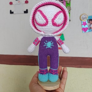 Puede incluir: Muñeca de ganchillo hecha a mano de una superheroína, con cabeza blanca y detalles rosas, y una capucha a juego. La muñeca lleva un mono morado con un emblema de araña turquesa y zapatos turquesa y rosas.