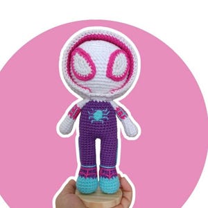 Może przedstawiać: Ręcznie robiona lalka amigurumi Spider-Gwen, z biało-różową maską, fioletowym kombinezonem i niebieskimi akcentami. Lalka jest szydełkowana i trzymana na drewnianej podstawie. Na zdjęciu widnieje napis "Spider Gwen" i "Patron de pago".