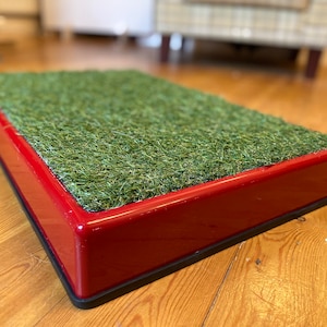 May include: Red plastic pet potty with a green artificial grass insert. The potty is on a wooden floor.