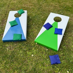 May include: Two cornhole boards with white, blue, and green painted sections. Each board has a hole and is surrounded by green and blue bean bags. The boards are on a grassy lawn.