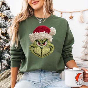 Könnte beinhalten: Grünes Sweatshirt mit einem gestickten Grinch-Gesicht mit Weihnachtsmütze. Das Gesicht des Grinch ist mit grünen, roten und weißen Fäden detailliert. Das Sweatshirt ist einfarbig grün und hat einen Rundhalsausschnitt.