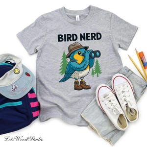 Puede incluir: Una camiseta gris con el texto "BIRD NERD" y un pájaro de dibujos animados con sombrero y prismáticos. El pájaro es azul y amarillo, con botas marrones. Zapatillas blancas, una mochila y un sombrero están junto a la camiseta.
