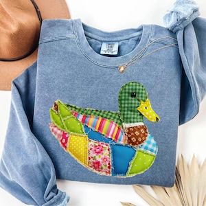 May include: A light blue sweatshirt with a patchwork duck design. The duck features a green checkered head, a yellow beak, and colorful wings with floral, striped, and polka dot patterns. A gold necklace with a pendant is visible.