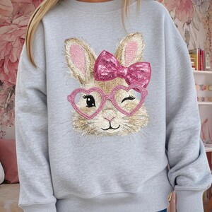 Puede incluir: Sudadera gris claro con un rostro de conejo bordado, gafas rosas en forma de corazón y un lazo de lentejuelas. El conejo está guiñando un ojo. La sudadera es de estilo casual y cómodo.