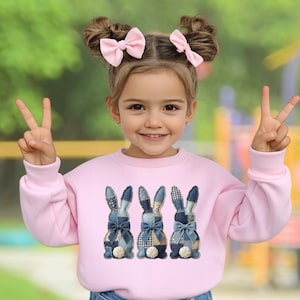 Puede incluir: Una sudadera rosa claro con tres diseños de conejos de mezclilla con lazos azules y detalles blancos. La niña lleva lazos rosas en el pelo y hace el signo de la paz.