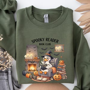 Puede incluir: Sudadera verde oliva con el texto "Spooky Reader Book Club". El gráfico presenta un pato de dibujos animados con un sombrero de bruja leyendo un libro junto a una chimenea con calabazas y murciélagos. Un diseño acogedor para Halloween.