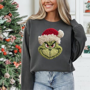 Puede incluir: Sudadera gris oscuro con un bordado de la cara del Grinch con un gorro de Papá Noel rojo y blanco. La cara del Grinch es verde con ojos rojos y una sonrisa traviesa. La sudadera tiene mangas largas y cuello redondo.