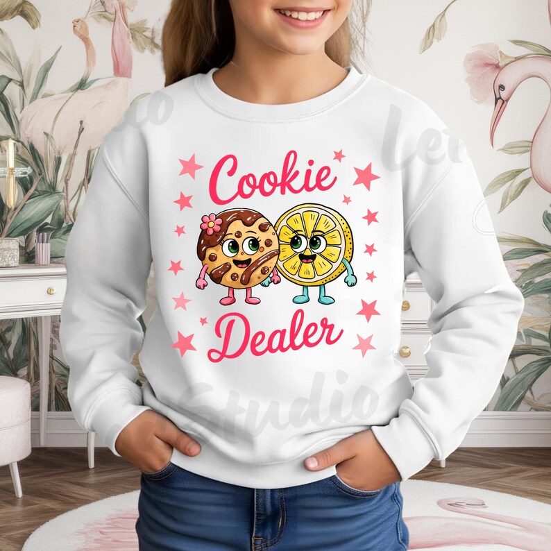 May include: White sweatshirt with the text "Cookie Dealer" in pink, featuring cartoon illustrations of a cookie and a lemon slice. The cookie has a flower and the lemon has a smiling face. Pink stars surround the text and images.