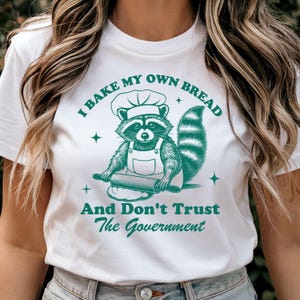 Puede incluir: Camiseta blanca con un gráfico verde de un mapache con un gorro de chef y extendiendo masa. El texto dice "Hago mi propio pan y no confío en el gobierno."
