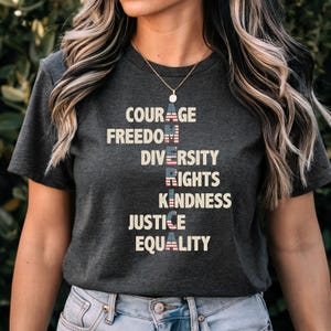 Puede incluir: Camiseta gris oscuro con las palabras "COURAGE, FREEDOM, DIVERSITY, RIGHTS, KINDNESS, JUSTICE, EQUALITY" en blanco. Las letras "GE", "DOM", "SITY", "GHTS", "NESS", "TICE" y "LITY" están diseñadas con un patrón de bandera estadounidense.