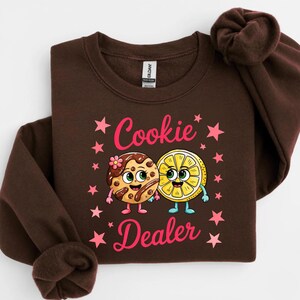 Sweatshirt featuring a cute cartoon cookie and lemon characters holding hands with the text “Cookie Dealer,” shown in a playful, colorful design; the design is available on t-shirts and sweatshirts in multiple sizes and a variety of colors.