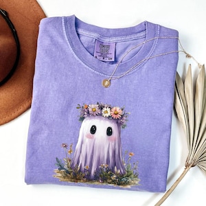 Puede incluir: Una camiseta Comfort Colors lavanda con un lindo fantasma que lleva una corona de flores. El fantasma tiene grandes ojos negros y mejillas rosadas. Un collar de oro con un colgante descansa sobre la camiseta. La camiseta está doblada y lista para usar.
