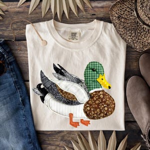 Puede incluir: Camiseta color crema con una aplicación de pato. El pato tiene una cabeza a cuadros verdes, un cuerpo con estampado floral marrón y alas grises y blancas. La camiseta se combina con un collar, jeans, un sombrero y zapatos.