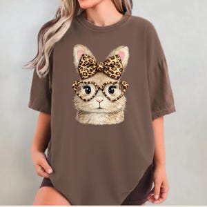 Puede incluir: Camiseta marrón con un diseño de cara de conejo. El conejo lleva gafas en forma de corazón y un lazo con estampado de leopardo. La camiseta es de color marrón liso y tiene un aspecto suave y texturizado.