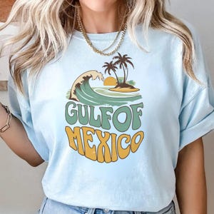 Puede incluir: Una camiseta azul claro con un diseño gráfico retro que presenta una ola, palmeras y el texto "GULF OF MEXICO".