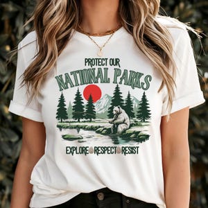 Puede incluir: Camiseta blanca con un diseño gráfico de un oso pescando en un lago con montañas y un sol rojo en el fondo. El texto "Protect Our National Parks" está por encima del oso y el texto "Explore Respect Resist" está por debajo del oso.