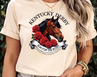 May include: A beige t-shirt with a graphic of a brown horse's head with a black mane and bridle. The graphic is surrounded by red roses and a banner that reads "Kentucky Derby - Run for the Roses - Churchill Downs, Louisville, Kentucky".