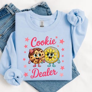Sweatshirt featuring a cute cartoon cookie and lemon characters holding hands with the text “Cookie Dealer,” shown in a playful, colorful design; the design is available on t-shirts and sweatshirts in multiple sizes and a variety of colors.