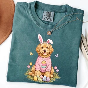 Puede incluir: Camiseta Comfort Colors verde azulado con un perro de dibujos animados con orejas de conejo y un suéter rosa con un diseño de huevo de Pascua. El perro está sentado en la hierba con huevos coloridos y mariposas. Un collar dorado está sobre la camisa.