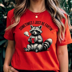 Puede incluir: Camiseta roja con un mapache sosteniendo una taza de café y el texto "Sometimes I Just Be Fartin'" impreso en ella.