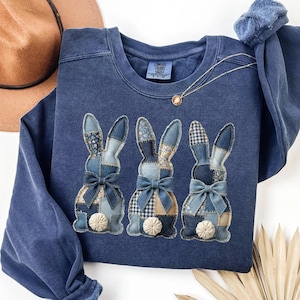 Può includere: Una felpa blu navy con tre coniglietti applicati con motivi patchwork e fiocchi. I coniglietti hanno diversi motivi, tra cui floreali e gingham, nei toni del blu e del beige. La felpa ha girocollo e maniche lunghe.