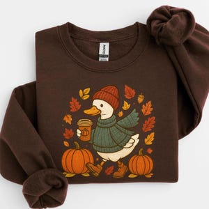 Puede incluir: Una sudadera marrón con un diseño de pato de dibujos animados. El pato lleva gorro, bufanda y botas, y sostiene una taza de café. El diseño incluye calabazas, hojas de otoño y bellotas. La sudadera es de color marrón liso.