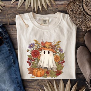 Puede incluir: Camiseta de color crema con un fantasma caprichoso que lleva un sombrero marrón adornado con flores y hojas de otoño. Una calabaza se encuentra debajo del fantasma, rodeada de flores coloridas. La camiseta es de la marca Comfort Colors.