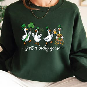 Puede incluir: Sudadera verde oscuro con un diseño del Día de San Patricio. Muestra cuatro gansos de dibujos animados con sombreros verdes, sosteniendo un trébol, cerveza y una bolsa de monedas de oro. El texto "just a lucky goose" está debajo.