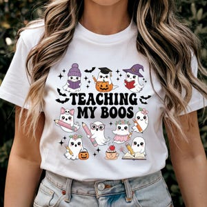Puede incluir: Camiseta blanca con un gráfico colorido que presenta fantasmas de dibujos animados con varios atuendos, incluyendo un sombrero de bruja y una gorra de graduación. El texto "TEACHING MY BOOS" está impreso en negro. El diseño incluye murciélagos, estrellas y calabazas.