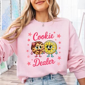 Sweatshirt featuring a cute cartoon cookie and lemon characters holding hands with the text “Cookie Dealer,” shown in a playful, colorful design; the design is available on t-shirts and sweatshirts in multiple sizes and a variety of colors.