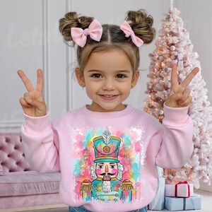 Puede incluir: Una sudadera rosa claro con un diseño de acuarela de un cascanueces. El cascanueces lleva un uniforme azul y dorado y un sombrero alto. El fondo tiene salpicaduras de rosa, azul y amarillo. La persona sonríe y lleva lazos rosas.