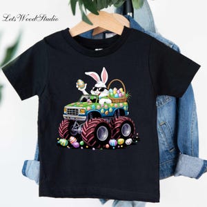May include: A black t-shirt featuring a green monster truck with a white bunny rabbit wearing sunglasses and holding an Easter egg. The truck is carrying a basket of Easter eggs. The truck has large tyres and is surrounded by Easter eggs.