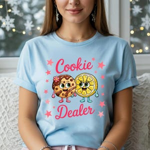 Sweatshirt featuring a cute cartoon cookie and lemon characters holding hands with the text “Cookie Dealer,” shown in a playful, colorful design; the design is available on t-shirts and sweatshirts in multiple sizes and a variety of colors.