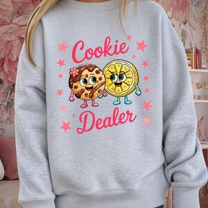 May include: A light gray sweatshirt featuring the text "Cookie Dealer" in pink, with pink stars. The graphic includes a cartoon cookie and a lemon slice, both with smiling faces and arms and legs.