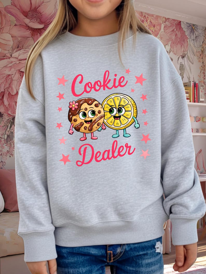 May include: A light gray sweatshirt featuring the text "Cookie Dealer" in pink, with pink stars. The graphic includes a cartoon cookie and a lemon slice, both with smiling faces and arms and legs.